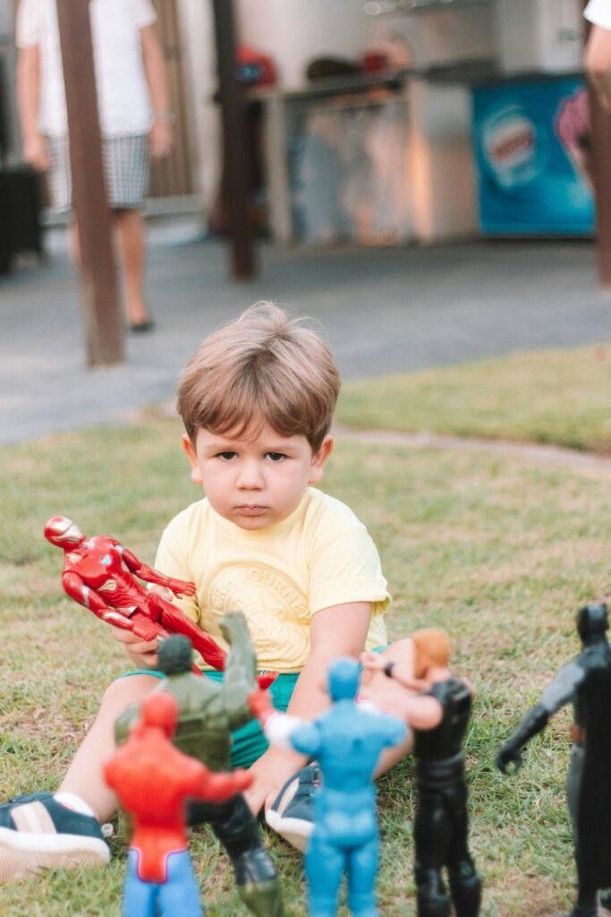 A young boy sitting on grass playing with superhero action figures on a sunny day.